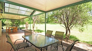 Een terras buiten met een glazen tafel en stoelen, omgeven door bomen en een gazon.