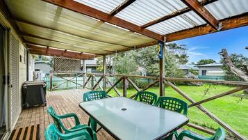 A covered outdoor area with a table and chairs, a wooden deck, and a view of the surrounding greenery.