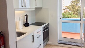 Une cuisine de taille réduite, avec des meubles blancs, une cuisinière et un évier. Une petite fenêtre offre une vue sur un immeuble et des arbres.
