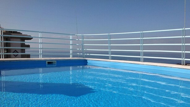 Une piscine d’un bleu éclatant, bordée d’une rambarde en métal, un petit bâtiment sur la gauche et un ciel dégagé en arrière-plan.