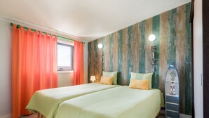 A bedroom with two beds, orange curtains, a window, a lamp, and a shelf with decorative items. - HC Torreira Beach (Torreira Aveiro)