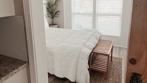 A bedroom with a bed, a small wooden bench, a potted plant, and a window with blinds.