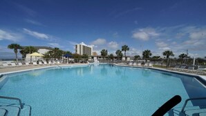 A large outdoor swimming pool with lounge chairs and umbrellas, surrounded by palm trees and a building in the background.