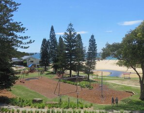A park with playground equipment, a building, and a beach in the background.