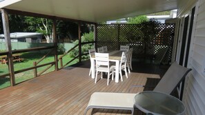 Un patio couvert avec une terrasse en bois, des meubles d’extérieur blancs et une vue sur une cour arrière agrémentée d’une clôture et de verdure.
