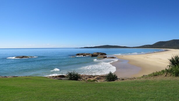 A coastal landscape with a sandy beach, clear blue water, and rocky outcrops. - MRS MOTTEES COTTAGE - SHORT STROLL TO BEACH, AIRCONDITIONED & PET FRIENDLY. (South West Rocks)