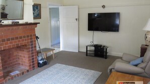 A living room with a brick fireplace, a flat-screen TV mounted on the wall, a sofa, and a small table with a lamp. - MRS MOTTEES COTTAGE - SHORT STROLL TO BEACH, AIRCONDITIONED & PET FRIENDLY. (South West Rocks)