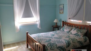 A bedroom with a bed, a nightstand, a lamp, and a window with curtains. - MRS MOTTEES COTTAGE - SHORT STROLL TO BEACH, AIRCONDITIONED & PET FRIENDLY. (South West Rocks)