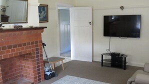 A living room with a brick fireplace, a flat-screen TV mounted on the wall, a sofa, and a small table with a lamp. - MRS MOTTEES COTTAGE - SHORT STROLL TO BEACH, AIRCONDITIONED & PET FRIENDLY. (South West Rocks)