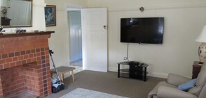 A living room with a brick fireplace, a flat-screen TV mounted on the wall, a sofa, and a small table with a lamp. - MRS MOTTEES COTTAGE - SHORT STROLL TO BEACH, AIRCONDITIONED & PET FRIENDLY. (South West Rocks)