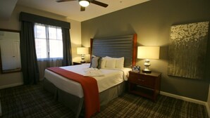 A hotel room with a bed, two bedside tables, a ceiling fan, a window with curtains, and a wall-mounted artwork.