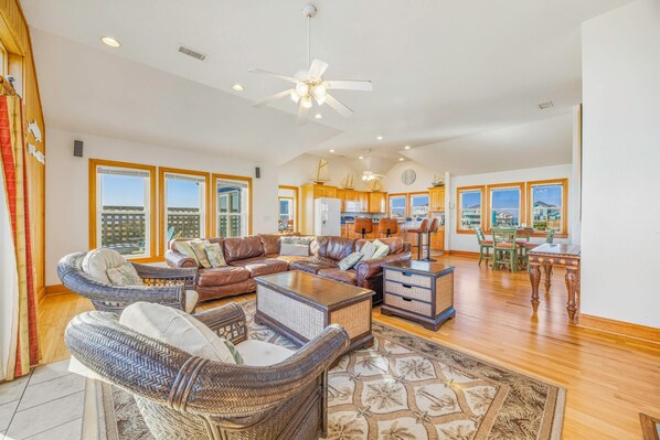 A spacious living room with a large sofa, a coffee table, and wicker chairs. There is a kitchen area with wooden cabinets and a dining table with chairs. - Oceanfront Home w/ Private Pool, Hot Tub, Elevator, Game Room, Home Theater (Frisco)