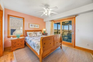 A bedroom with a wooden bed, a nightstand, a lamp, a framed picture, and a view of the outdoors through a sliding glass door. - Oceanfront Home w/ Private Pool, Hot Tub, Elevator, Game Room, Home Theater (Frisco)