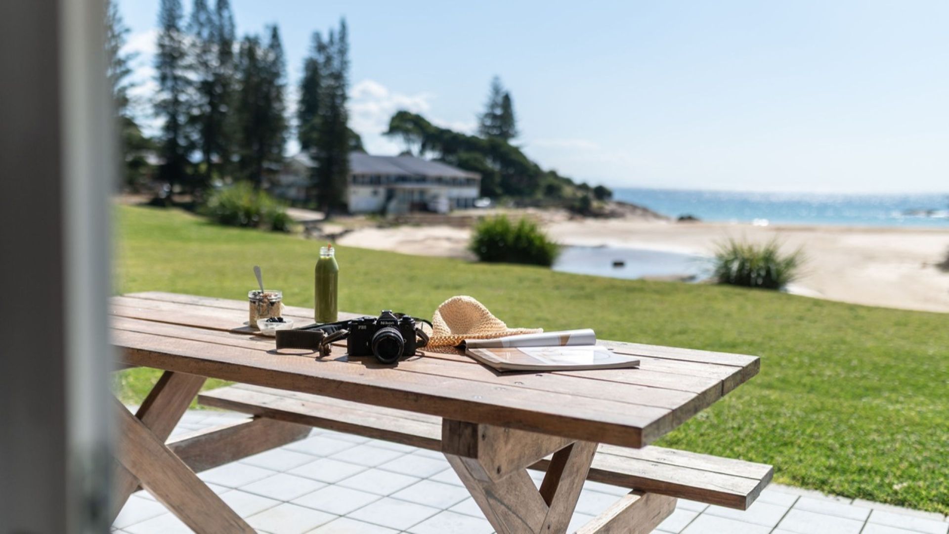 A wooden picnic table with a camera, a straw hat, a bottle, and a jar of snacks.