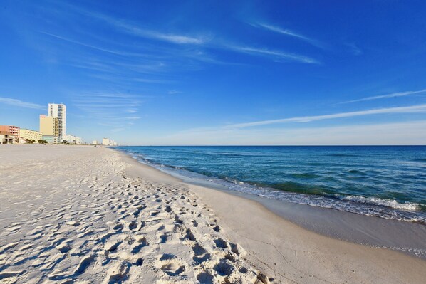 A sandy beach with footprints leading to the water, a clear blue sky, and buildings in the background.