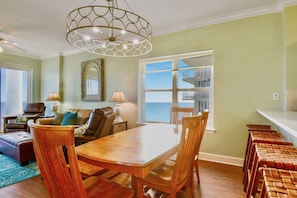 A dining area with a wooden table and chairs, a chandelier, and a view of the ocean through a large window.