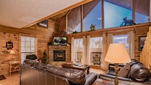 A cozy cabin interior with a stone fireplace, leather sofas, a television, and large windows offering a view of the outdoors. - ⛰️A Bird Song: Spectacular Views | Minutes from Pigeon Forge & Gatlinburg🌲 (Sevierville)