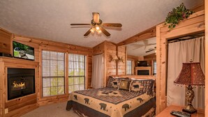 A cozy log cabin bedroom with a fireplace, a large bed with a bear-themed quilt, a ceiling fan, and a television mounted on the wall. - ⛰️A Bird Song: Spectacular Views | Minutes from Pigeon Forge & Gatlinburg🌲 (Sevierville)