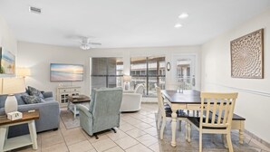 A living room with a sofa, armchair, dining table, and chairs. There is a TV, a lamp, and a decorative wall piece. - Family friendly, beach views! (Gulf Shores)