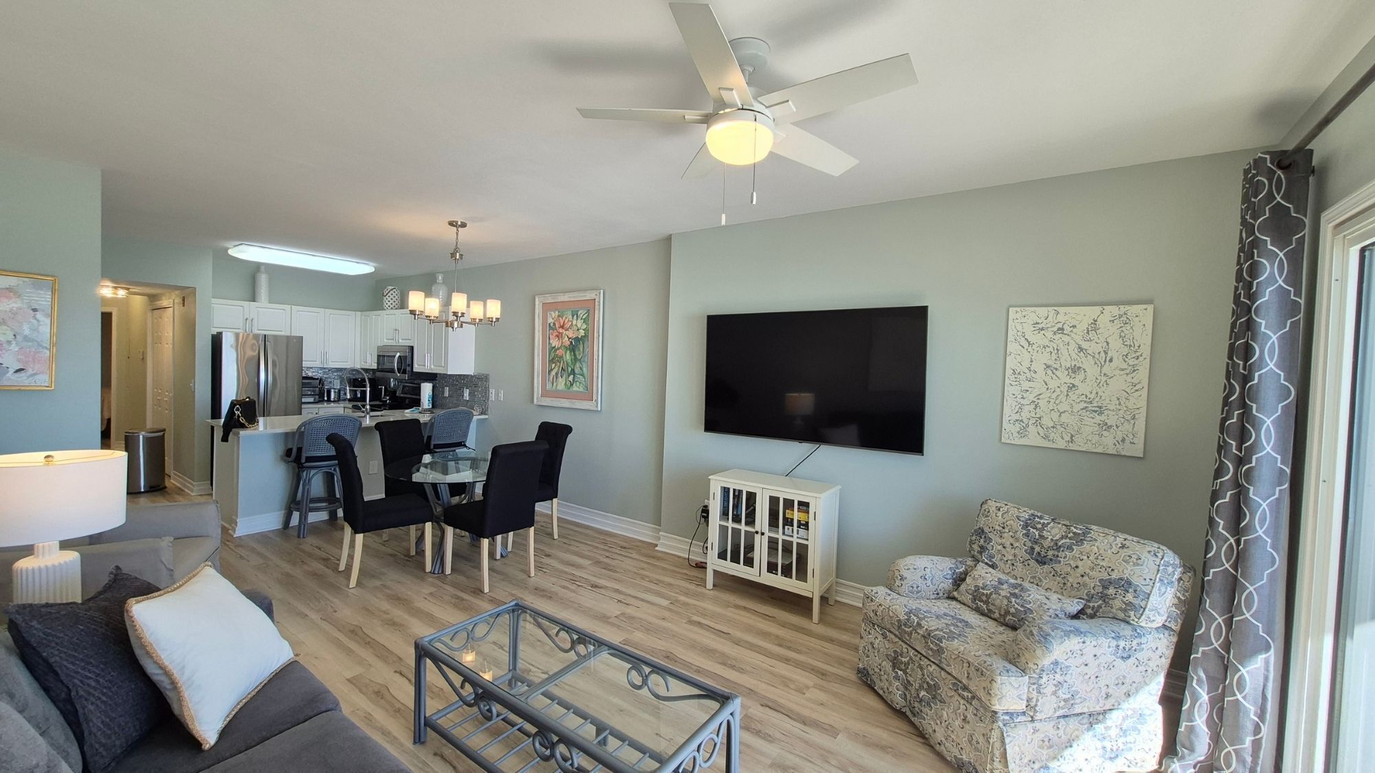 A modern living room with a sofa, coffee table, and a TV. There's a kitchen area with a dining set and a ceiling fan.