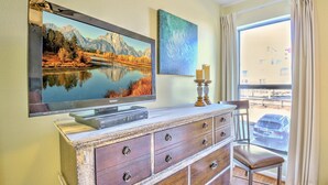 A room with a flat-screen TV mounted on the wall, a wooden dresser with drawers, a chair, a window with curtains, and a painting above the TV.