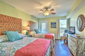 A room with two beds, a ceiling fan, a television, a dresser, and a window with a view of the outside.