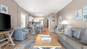 A spacious living room with a flat-screen TV, a sofa, a coffee table, and a dining area with chairs and a kitchen in the background. - Dog friendly beachfront condo with sunset view (Gulf Shores)