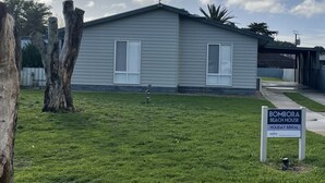 A single-story beach house with a covered porch, a small lawn, and a sign indicating it's available for holiday rental. - This large holiday home is fully equipped with everything you will need. (Robe)