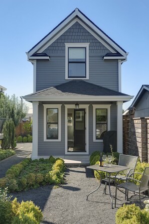 A two-story house with a front porch, a small garden with gravel and shrubs, and a metal table with chairs.