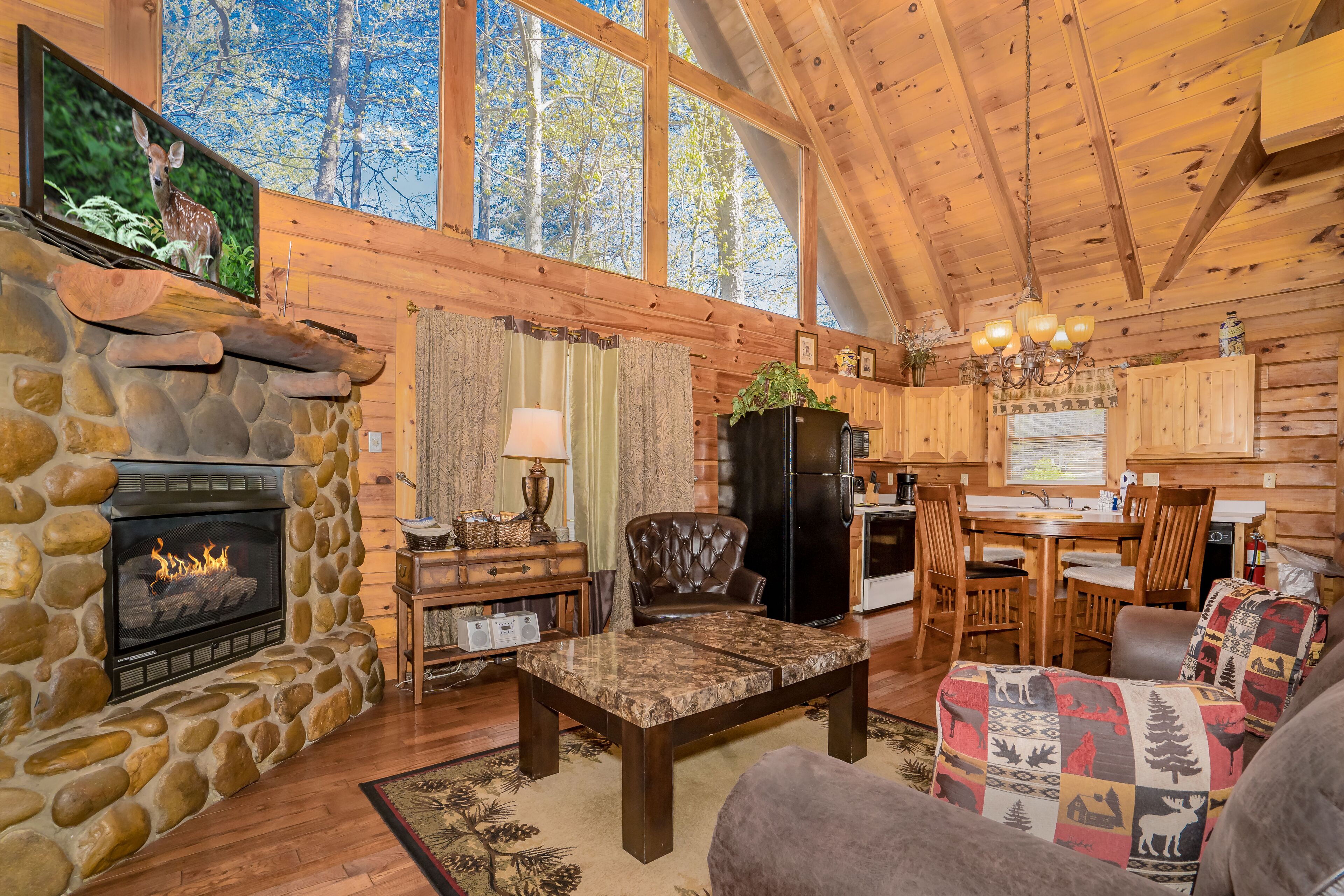A cozy cabin interior with a stone fireplace, wooden walls, a dining area, and a large window overlooking trees.