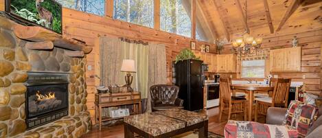 A cozy cabin interior with a stone fireplace, wooden walls, a dining area, and a large window overlooking trees.
