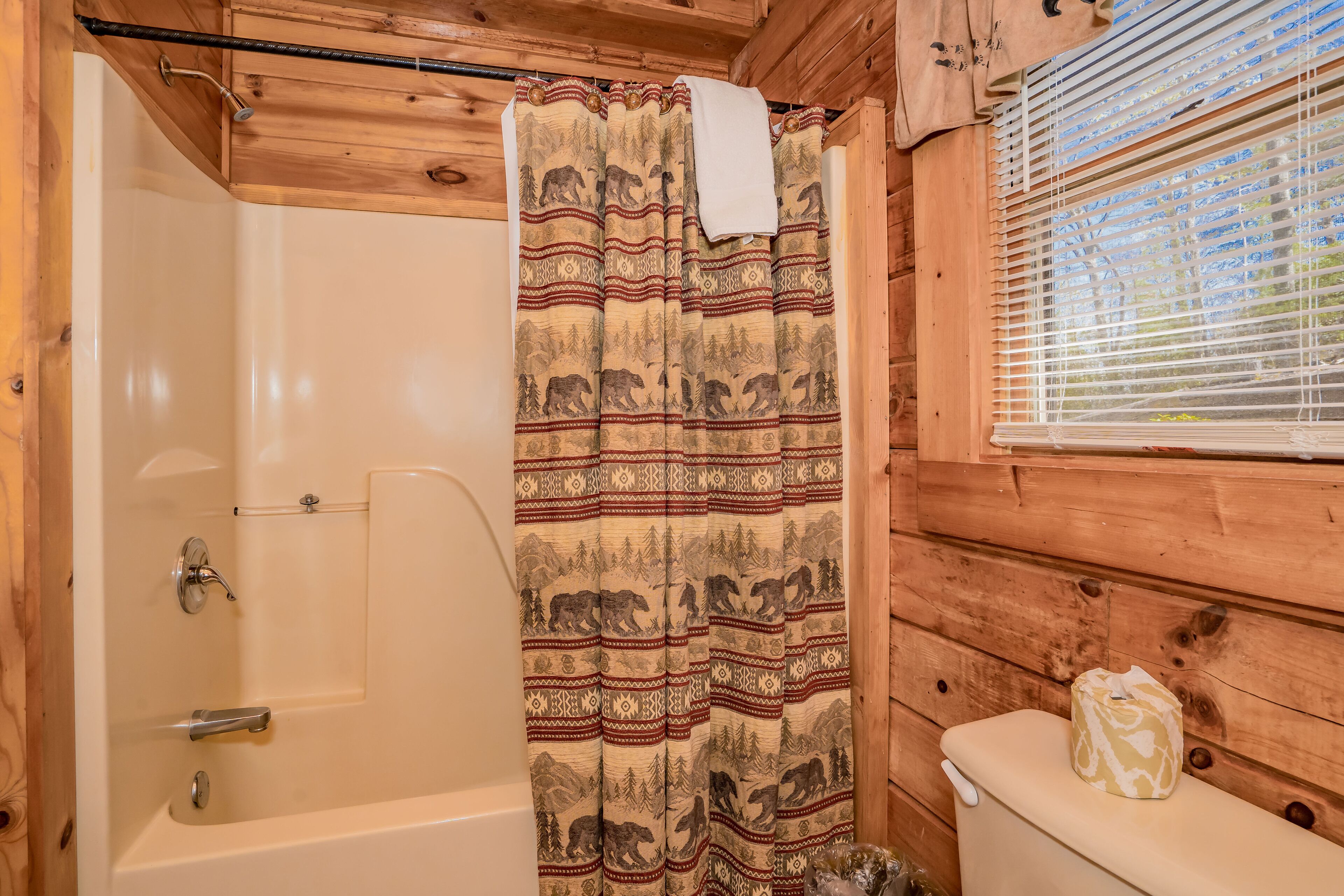 Une salle de bain rustique, avec des murs en bois, comprenant une baignoire, des toilettes et un rideau de douche orné de motifs animaliers.