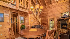 Interior de una cabaña de madera con comedor, chimenea y escalera.