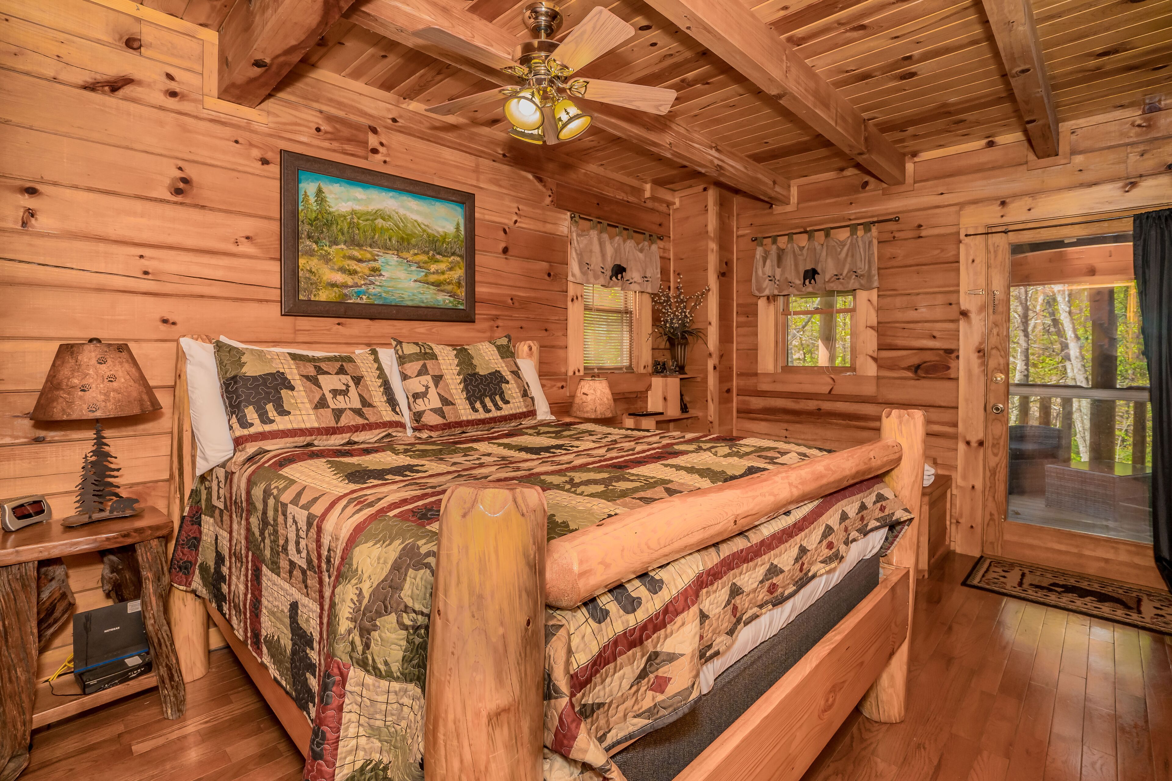Camera da letto in stile baita con un letto grande, una testiera in legno, una ventola a soffitto, un quadro raffigurante un paesaggio e una finestra con tende.