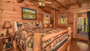 Un dormitorio tipo cabaña de troncos con una cama grande, cabecera de madera, ventilador de techo, un cuadro de un paisaje y una ventana con cortinas.
