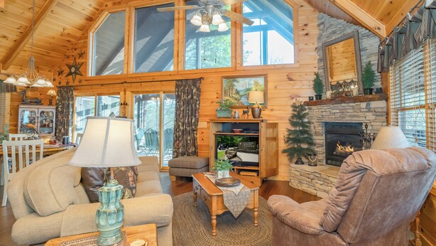 A cozy log cabin interior with a fireplace, wooden furniture, and a large window. - 🌲Night Moves Over The Smokies 🌙 Hot Tub, Pool Table & Porch Swing (Sevierville)