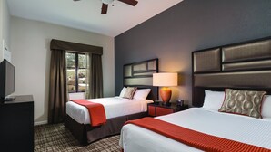 A hotel room with two beds, a television, a window with curtains, a ceiling fan, and a nightstand with a lamp.