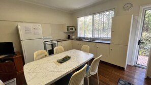 A compact kitchen with a dining table, chairs, a refrigerator, a microwave, and a window with blinds. - Cabenay Unit 2 - Close to Beach (South West Rocks)