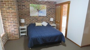 A bedroom with a brick wall, a bed with a blue bedspread, two bedside tables with lamps, and a door leading to another room. - BIRCHGROVE 3 - 400 METRES TO BEACH (SOUTH WEST ROCKS)