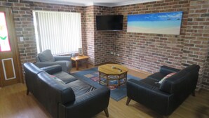A living room with brick walls, a flat-screen TV mounted above a painting, and two black leather sofas with cushions. - BIRCHGROVE 3 - 400 METRES TO BEACH (SOUTH WEST ROCKS)