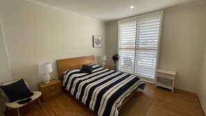 A bedroom with a bed, bedside tables, a chair, a standing fan, and a window with blinds. - TRADEWINDS UNIT 7 - BEACH VIEWS (South West Rocks)