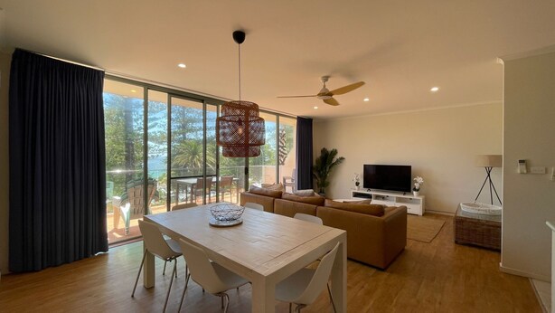 A modern living room with a dining area, large windows, a ceiling fan, and a flat-screen TV. - TRADEWINDS UNIT 7 - BEACH VIEWS (South West Rocks)