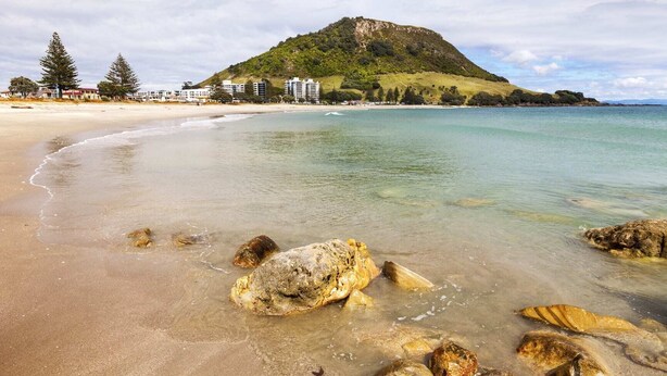 A beach with clear water, rocks, and a hill with buildings in the background. - Awesome Downtown Location - Park & Walk (Tauranga)