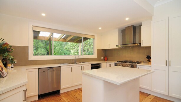 A modern kitchen with white cabinetry, a central island, a stainless steel dishwasher, and a window with a view of greenery.