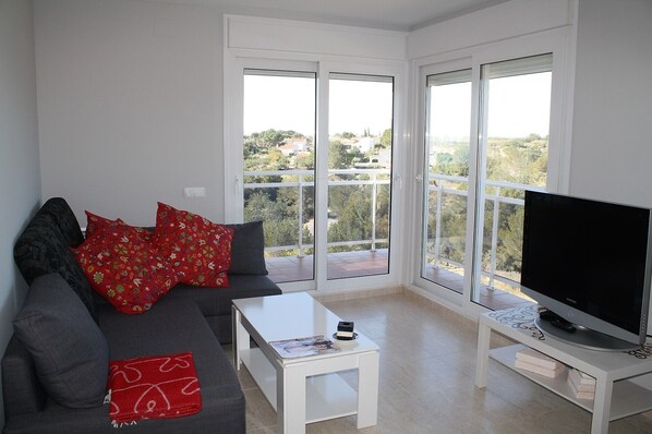 A modern living room with a grey sofa, red patterned pillows, a white coffee table, and a flat-screen TV on a white stand. - Beautiful apartment with sea views, 5 min walk to the beach. Cap roig (El Perelló)