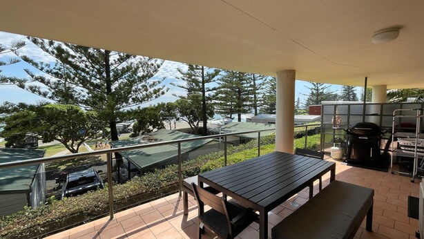 Un balcon avec une table et des chaises, donnant sur un parking et des arbres.