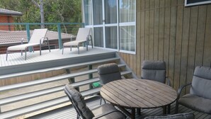 Una terraza de madera con mobiliario exterior, que incluye una mesa y sillas, y vistas a un edificio con grandes ventanales.
