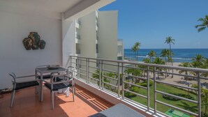 A balcony with a table and chairs, overlooking a beach with palm trees and a clear blue sky. - Trendy Beach Scene at a Harmony Beachfront Condo in Costa del Sol (Playa Juan Dolio)