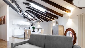 A modern living room with a grey sofa, wooden beams on the ceiling, a kitchen in the background, and a round mirror on the wall.