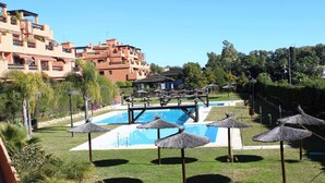 A pool area with a clear blue pool, surrounded by grass and palm trees. There are thatched umbrellas and a small bridge over the pool. - Ideal and front line beach location (Estepona)